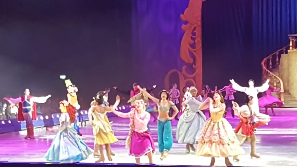 A group of performers in colourful costumes, including several Disney princesses, dance together on an ice rink stage with dramatic lighting. In the background, ornate golden decorations and a winding staircase are visible. The scene is lively and festive.