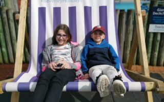 Two smiling children sit on a giant striped deckchair with "Beverley Holidays" written above them. The girl on the left wears glasses, a grey jacket, and black trousers. The boy on the right wears a cap, blue hoodie, and grey trousers. A pool and wooden fence are in the background.