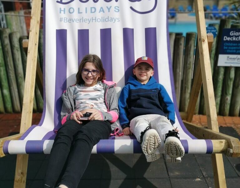 Two smiling children sit on a giant striped deckchair with "Beverley Holidays" written above them. The girl on the left wears glasses, a grey jacket, and black trousers. The boy on the right wears a cap, blue hoodie, and grey trousers. A pool and wooden fence are in the background.