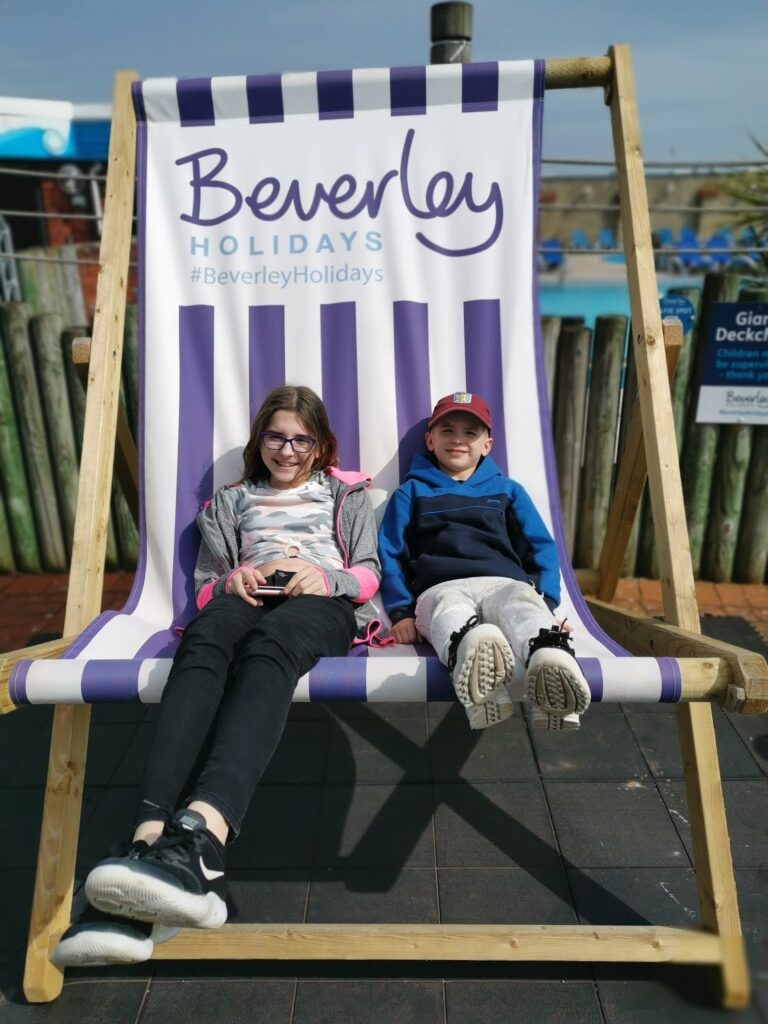 Two smiling children sit on a giant deckchair with purple and white stripes. The chair has a sign reading “Beverley HOLIDAYS #BeverleyHolidays.” There’s a pool and wooden fencing in the background. The kids look relaxed and happy.