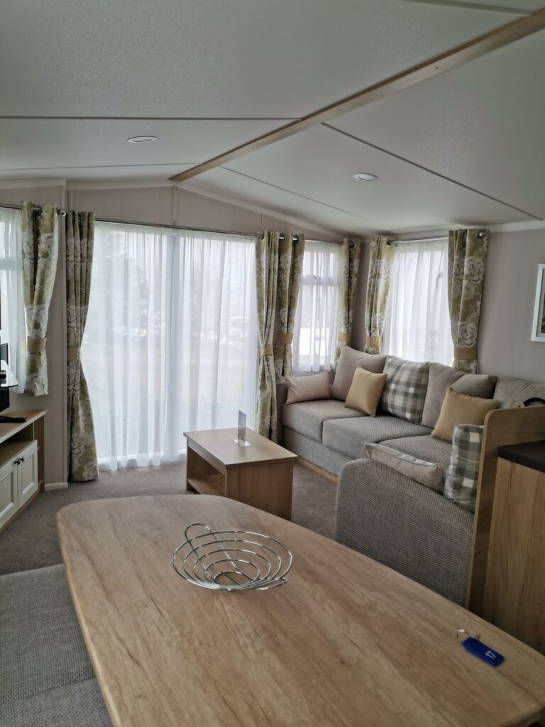 A bright living area with large windows covered by sheer curtains, a grey sofa with checked and beige cushions, a wooden coffee table, and a wooden dining table with a decorative metal bowl. Neutral tones and soft lighting create a cosy atmosphere.