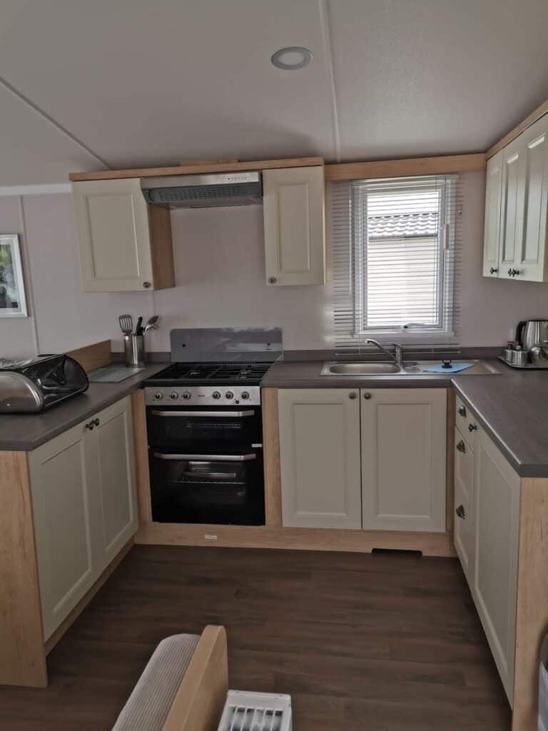 A modern, compact kitchen with light-coloured cupboards, a built-in oven and hob, a silver extractor hood, a toaster, utensils, kettle, and a microwave. A window above the sink lets in light, and the floor is wood-patterned.