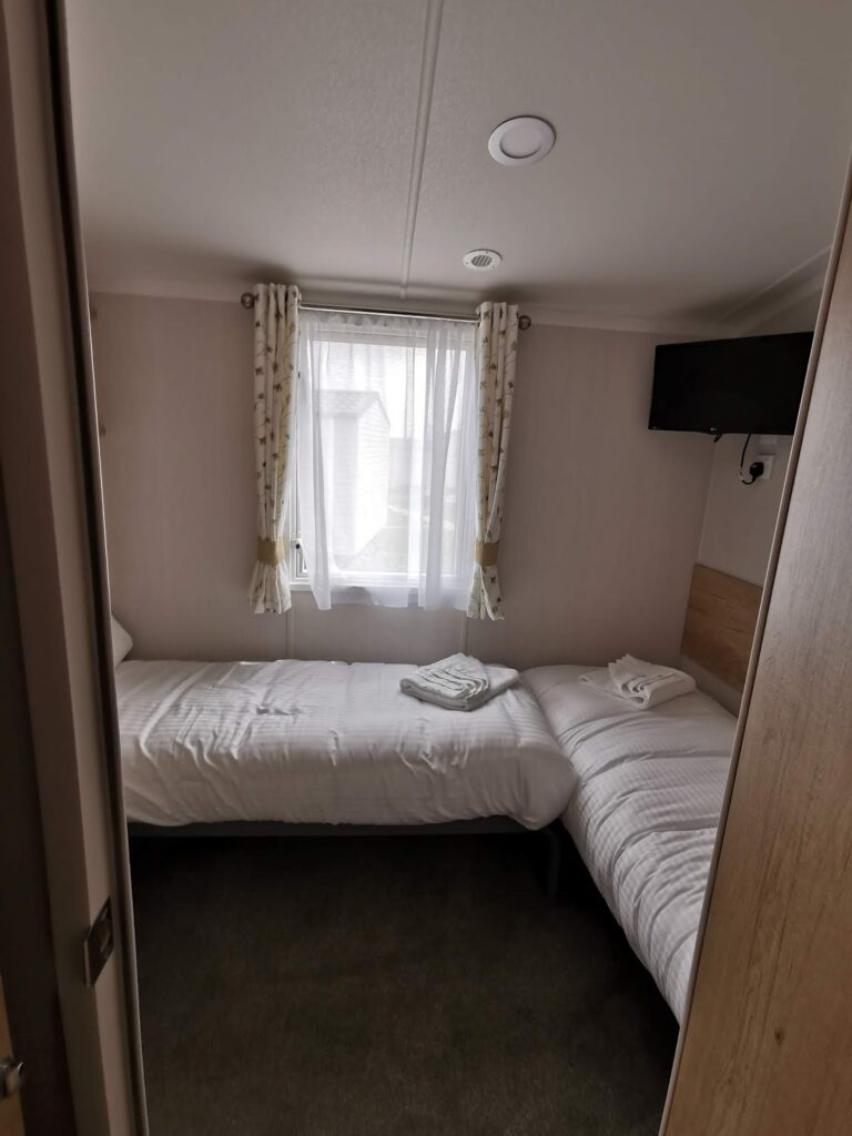 A small, tidy bedroom with two single beds along opposite walls, white bed linen, and folded towels on each bed. A window with patterned curtains lets in natural light. A wall-mounted TV is in the upper right corner, and the floor is carpeted.