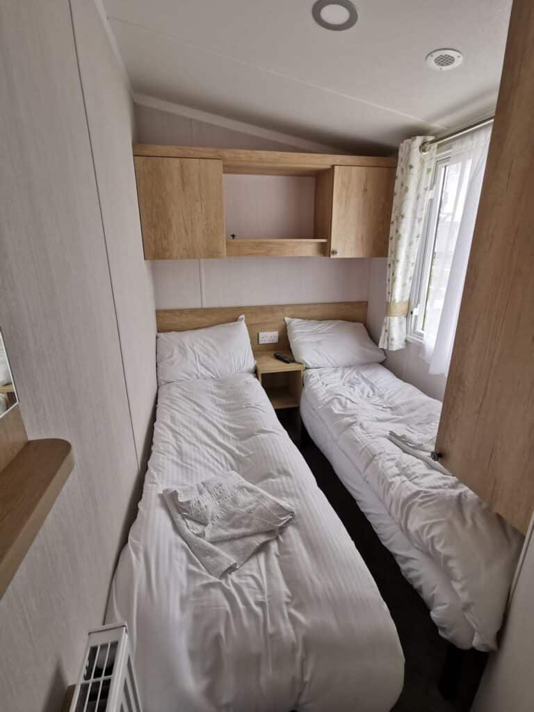 A small, narrow room with two single beds covered in white linen, separated by a wooden bedside table. There are shelves and cupboards above the beds, a window with light curtains on the right, and a folded towel on the left bed.