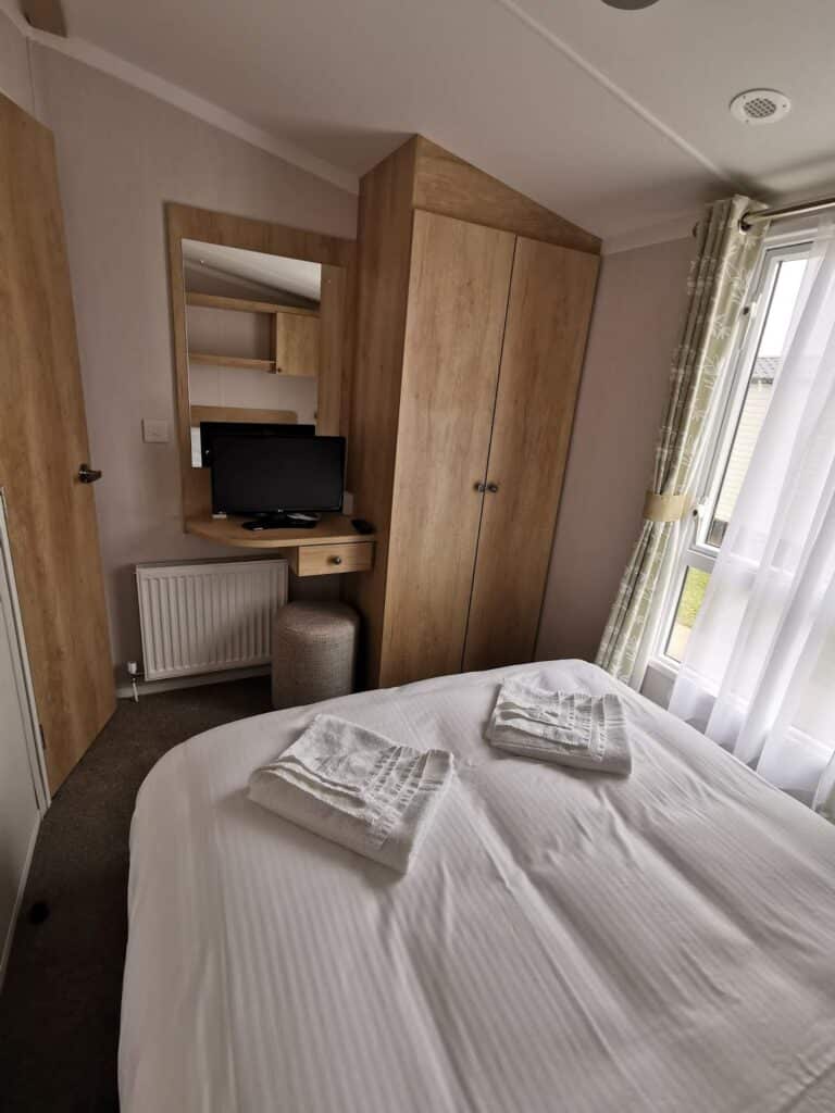 A cosy bedroom with a double bed covered in white linen and two folded towels on top. There’s a wooden wardrobe, a small desk with a TV, a cushioned stool, a mirror, and a window with sheer curtains letting in daylight.
