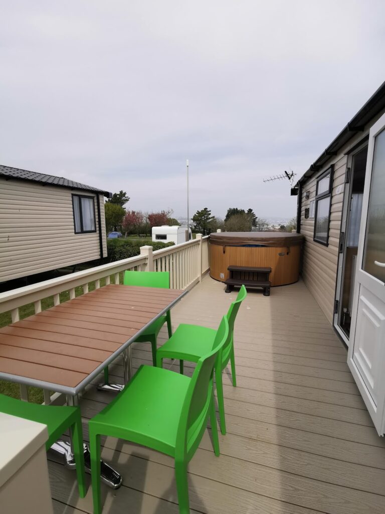 A decking with a brown table and three bright green chairs sits beside a tan caravan. At the far end, there is a round wooden hot tub. Another caravan and trees are visible in the background under an overcast sky.