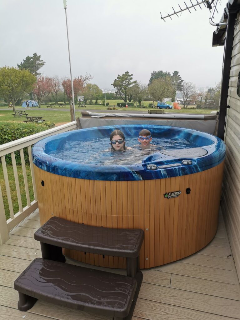 Two children wearing goggles relax in a blue and wooden round hot tub on a wooden decking. A small staircase leads up to the hot tub. The decking overlooks a grassy campsite with tents, trees, and people in the background under a cloudy sky.