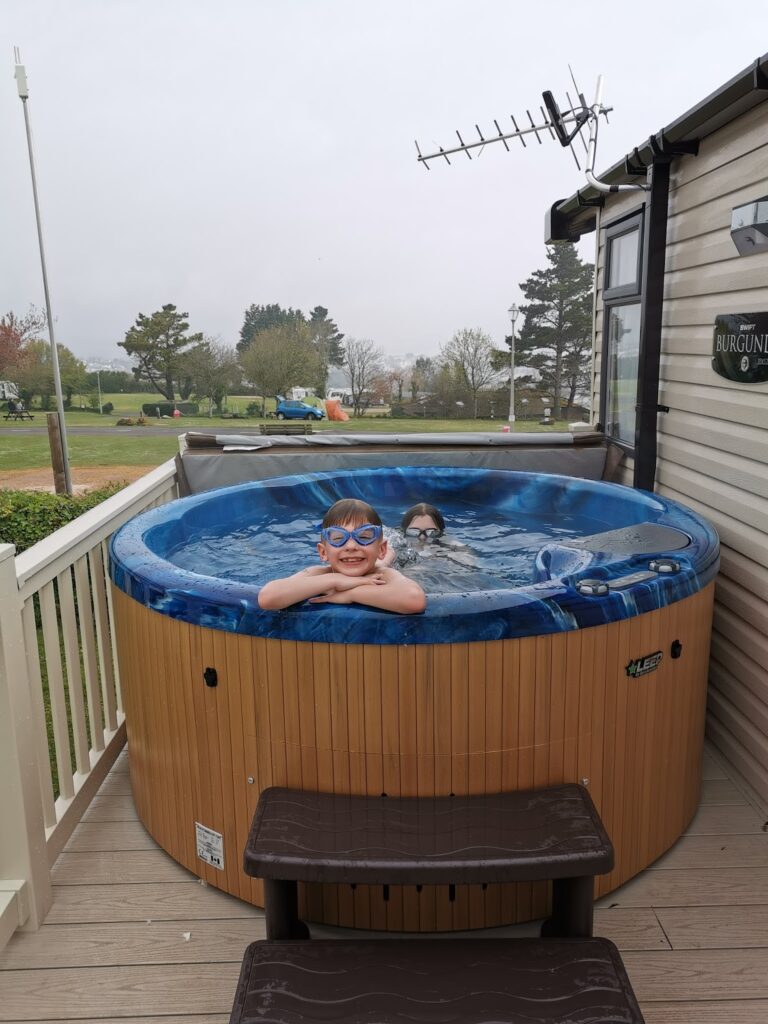 Two children are in a round outdoor hot tub on a decking. One child, wearing blue goggles, smiles at the camera, while the other is mostly submerged. There are trees, grass, and a caravan with an aerial in the background. The sky is overcast.