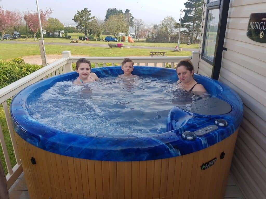 Three people, two children and one adult, are relaxing in a round blue hot tub on a wooden decking. The hot tub is next to a building, possibly a mobile home, with a grassy park and trees visible in the background on a sunny day.