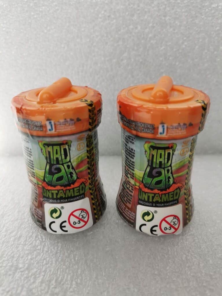 Two cylindrical Mad Lab Untamed toy containers with orange lids and straw-like tops stand side by side. The labels are colourful, displaying the logo, product name, and warning symbols including CE and a red “0-3” age warning. The background is light grey.