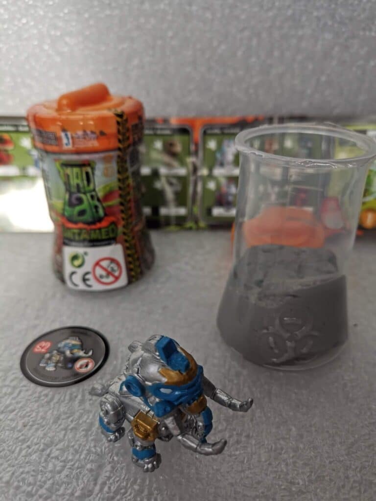 A small robot toy with blue and gold details stands on a grey surface. Nearby are a plastic beaker filled with grey slime, an orange-capped toy container labelled "Goo Lab Untamed," and a round token, with a toy instruction sheet blurred in the background.