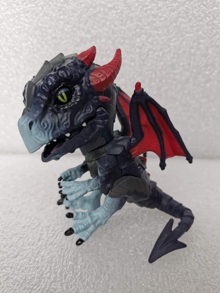 A toy dragon figure with blue and grey scales, red horns, green eyes, black claws, and red-and-black wings is posed against a textured white background. The dragon has detailed patterns on its face and a playful, fantasy-inspired design.