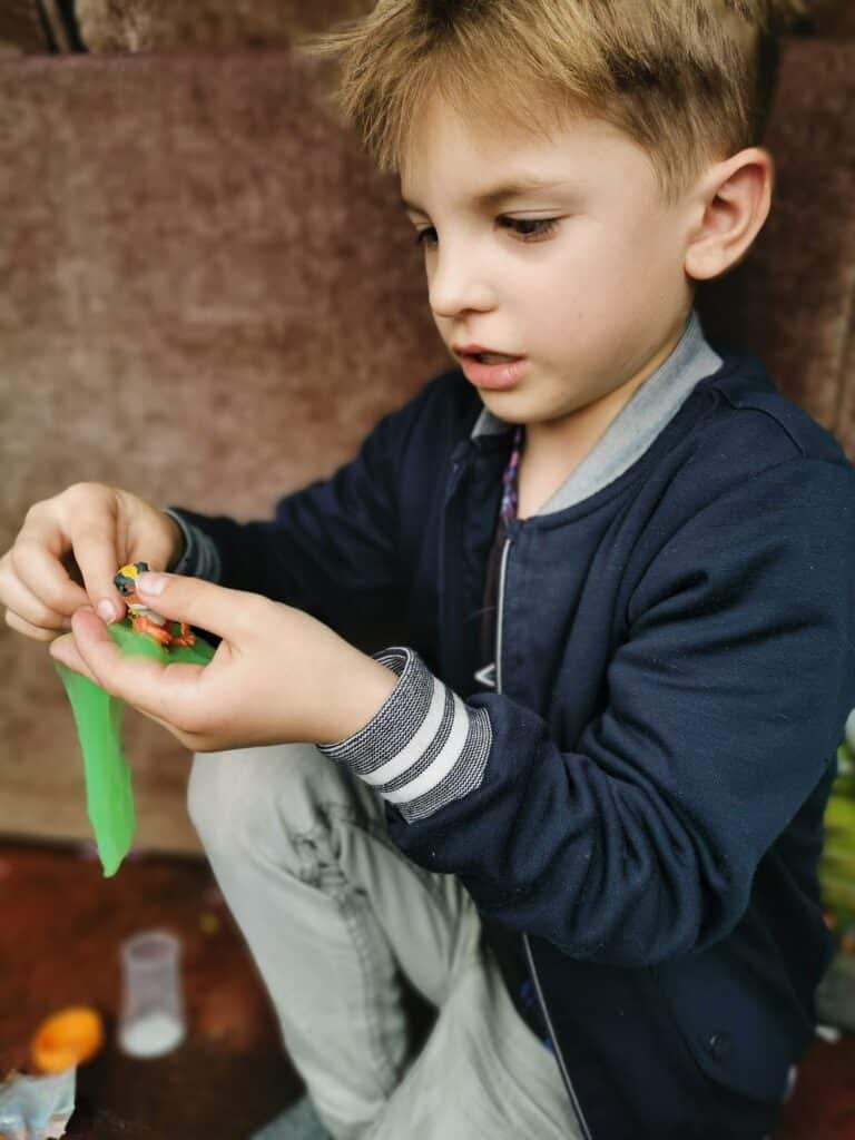 A young boy with light brown hair, wearing a navy jacket and grey trousers, sits and carefully stretches bright green slime between his hands. He looks focused on the activity. A small plastic container and orange lid are visible on the surface beside him.