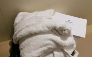A white bathrobe made of soft, fluffy towelling material is neatly folded on a beige bench. A white envelope with "The Battery" printed on it rests on top of the robe, and a dark folded towel is underneath. The background is a plain beige wall.