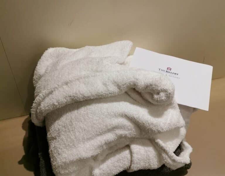 A white bathrobe made of soft, fluffy towelling material is neatly folded on a beige bench. A white envelope with "The Battery" printed on it rests on top of the robe, and a dark folded towel is underneath. The background is a plain beige wall.