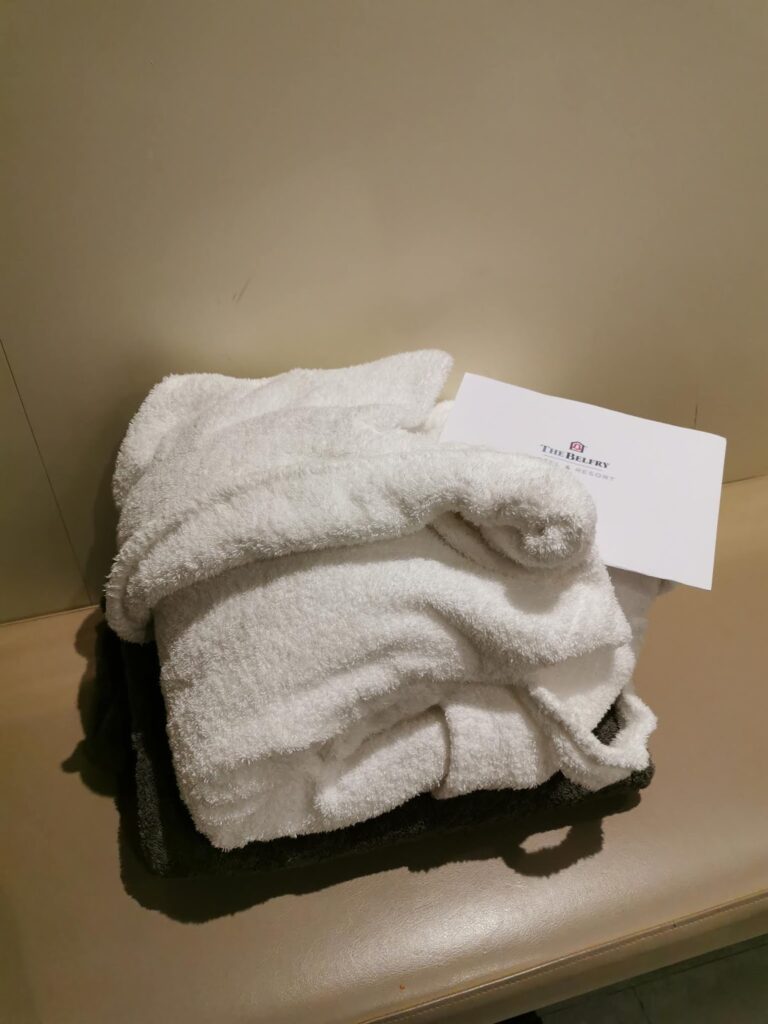 A neatly folded white dressing gown sits on a beige surface, with a white envelope labelled "The Ritz-Carlton" placed on top. A dark grey towel is partially visible underneath the dressing gown. The background is plain and neutral.