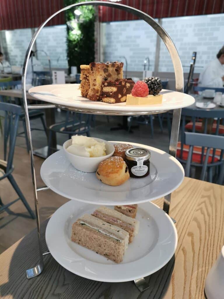 A three-tiered afternoon tea stand holds assorted desserts with berries on top, scones with clotted cream and jam, and finger sandwiches. The stand sits on a wooden table in a café, with chairs and blurred customers in the background.