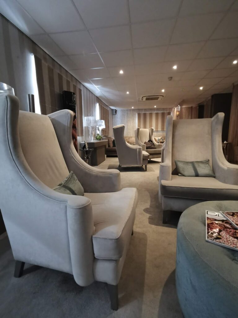 A modern lounge with light beige high-back armchairs, small decorative cushions, and a round ottoman with a magazine on top. The room features neutral tones, striped walls, soft lighting, and a suspended ceiling with recessed lights.