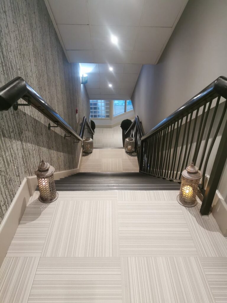 A tiled staircase with black handrails descends between two walls. Four decorative lanterns with lit candles are placed on the steps, two on each side. The stairwell leads to a landing with a window letting in daylight. The atmosphere is warm and inviting.