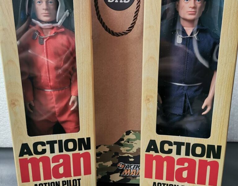 Two boxed Action Man dolls stand upright. The left box is labelled “Action Pilot” and contains a figure in a red jumpsuit with a white helmet. The right, “Action Sailor,” wears a navy uniform and cap. Behind them is a gift bag reading “#1 Dad”.