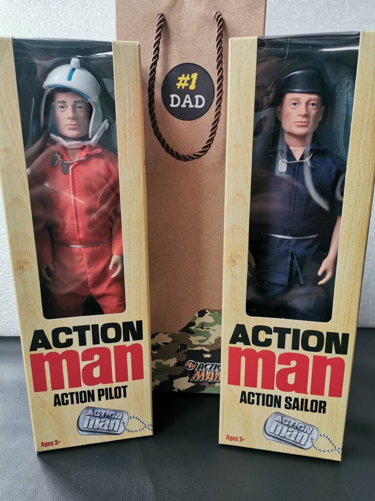 Two boxed Action Man dolls stand side by side: one wears a red pilot suit and white helmet, labelled "Action Pilot", and the other wears a navy sailor uniform and cap, labelled "Action Sailor". Between the boxes is a brown gift bag with a "#1 Dad" badge.