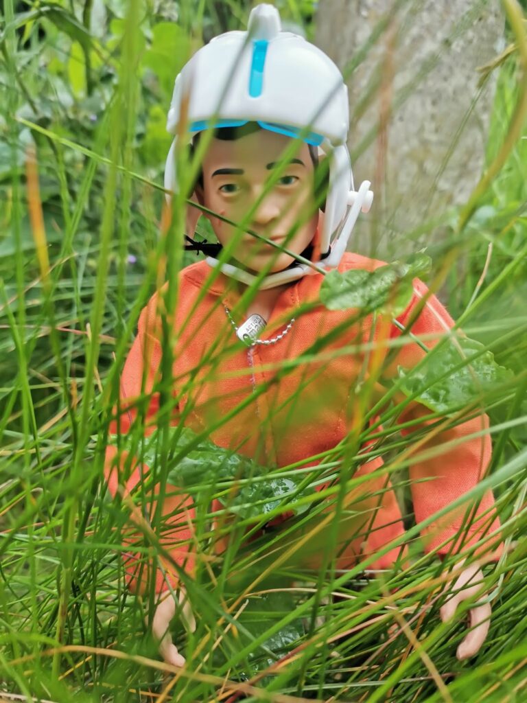 A plastic action figure in an orange outfit and white helmet is kneeling amid tall, green grass and leaves. The figure's facial features are visible through the blades of grass, and the background is blurred with more greenery and a stone surface.