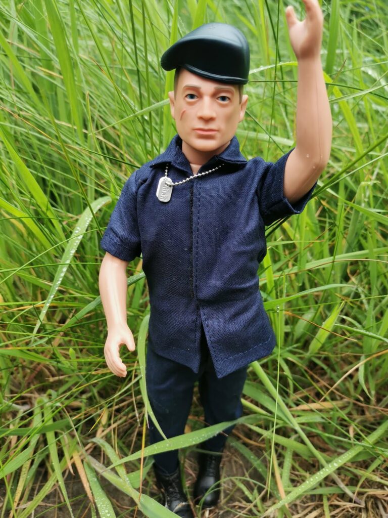 A toy soldier doll dressed in a navy blue uniform, black boots, and a black cap stands upright in tall green grass. The doll raises its right arm and wears a small silver identity tag necklace. The background is dense with vivid green blades of grass.