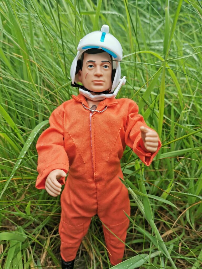 A male action figure in an orange jumpsuit and white helmet stands upright in tall, green grass. The figure has short dark hair, a serious facial expression, and its right hand raised slightly. The grass appears fresh, with some dew drops visible.