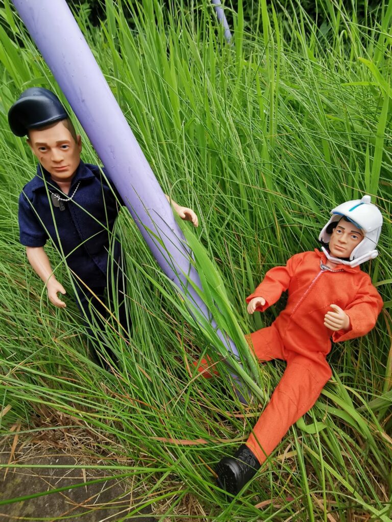 Two action figures are posed in tall grass outdoors. One wears a dark blue outfit and cap, standing against a purple pole. The other wears an orange jumpsuit and white helmet, sitting on the ground. Both appear to be in a rescue or emergency scene.