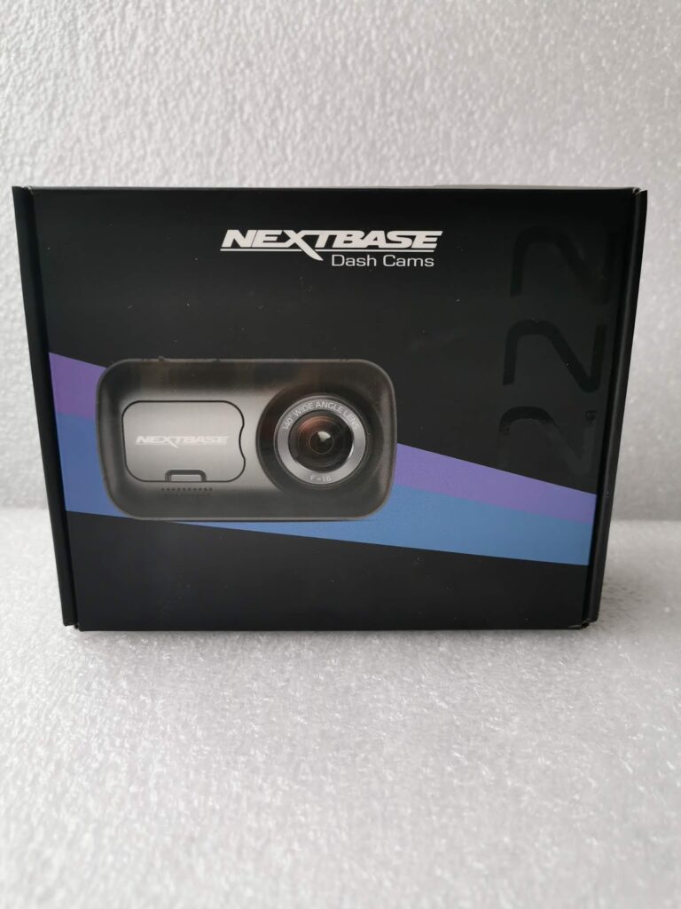 A black box of a Nextbase dash cam is shown against a light textured background. The packaging displays the dash cam image with the lens visible and includes the Nextbase Dash Cams logo at the top. Blue and purple diagonal stripes cross the front.