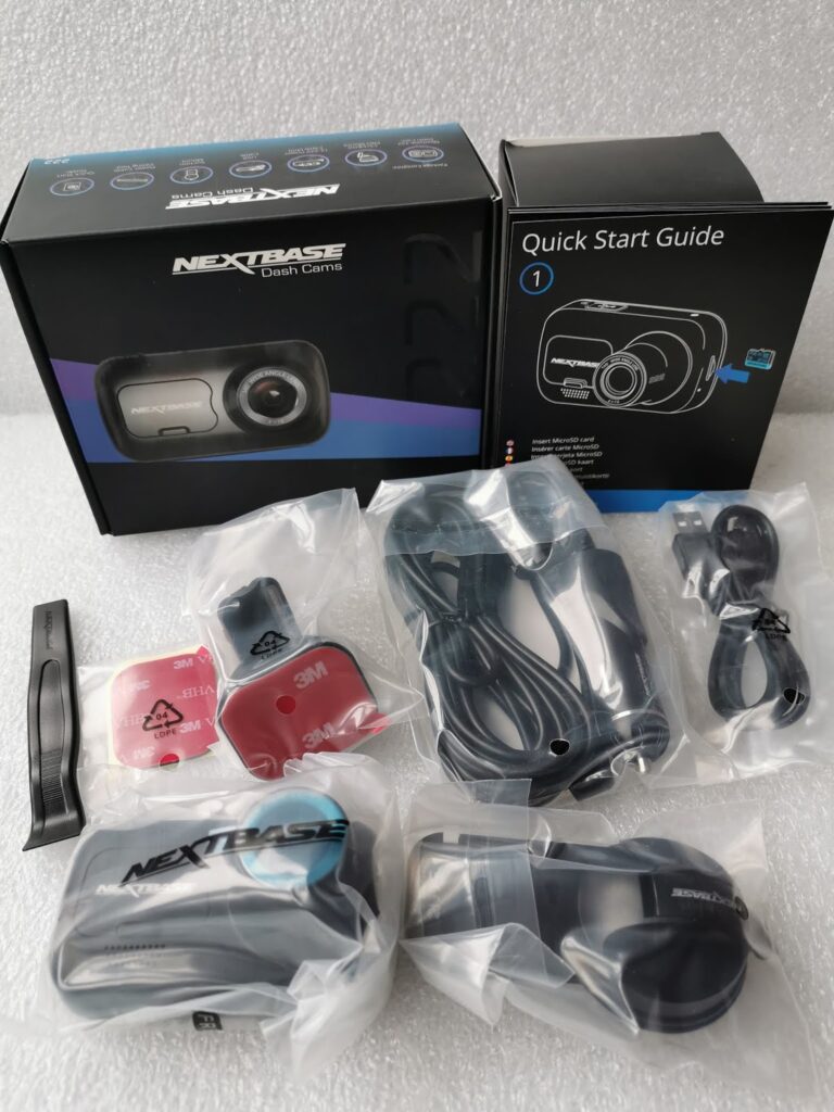 A Nextbase dash cam kit is displayed, including the dash cam, cables, mounting accessories, quick start guide, adhesive pads, and a black plastic tool, all neatly arranged on a white surface with some items in plastic packaging.