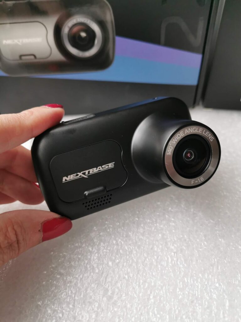A hand holds a black Nextbase dash cam with a large lens labelled “140° wide angle lens F=1.6.” The device has the Nextbase logo and a speaker grille on the front. A product box with a similar image is blurred in the background.