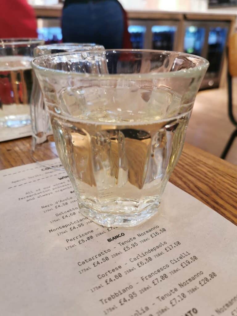 A clear glass filled with white wine sits on a wooden table atop a printed wine menu. Another glass, partially visible, is in the background along with blurred chairs, a person in blue jeans, and a counter with bottles in a softly lit setting.
