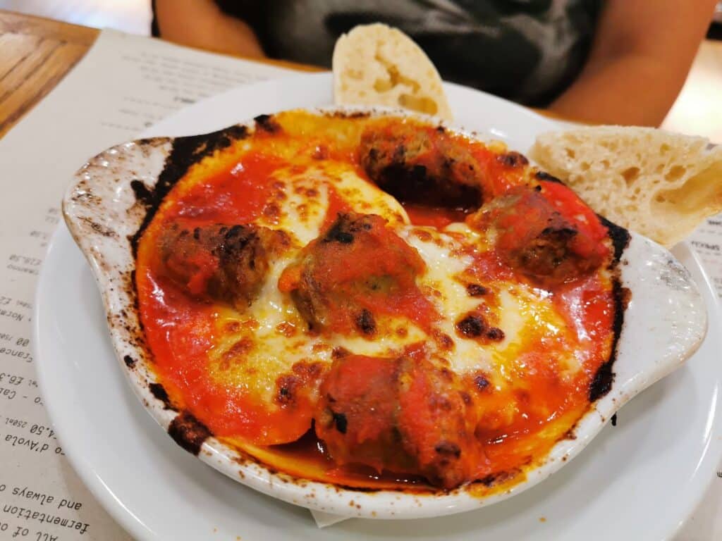 A white ceramic dish filled with baked meatballs in tomato sauce, topped with melted cheese and browned edges. Two pieces of crusty bread rest on the side, and the dish sits on a white plate with a printed menu partially visible underneath.