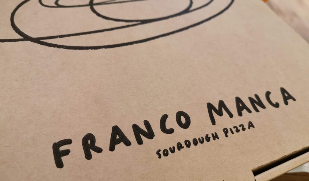 A close-up of a brown pizza box with a black spiral design and bold black text reading "FRANCO MANCA" above smaller text that says "Sourdough Pizza." Part of a drink is visible in the blurred background.