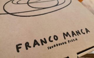 A close-up of a brown pizza box with a black spiral design and bold black text reading "FRANCO MANCA" above smaller text that says "Sourdough Pizza." Part of a drink is visible in the blurred background.