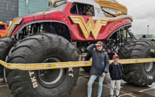 Two children stand in front of a large red Wonder Woman monster lorry with oversized tyres, parked outside a stadium with a "RICOH" sign. The lorry is behind yellow barrier tape, and the day appears cloudy and wet.