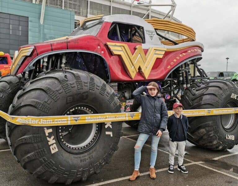 Two children stand in front of a large red Wonder Woman monster lorry with oversized tyres, parked outside a stadium with a "RICOH" sign. The lorry is behind yellow barrier tape, and the day appears cloudy and wet.