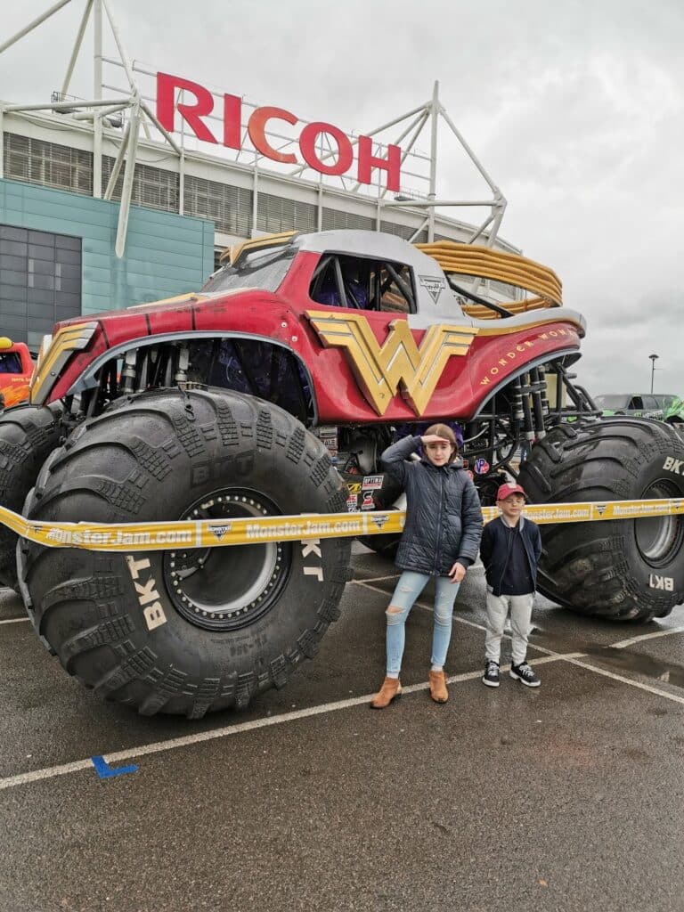 Two children pose in front of a large Wonder Woman monster truck with massive tyres, parked outside a building with a red "RICOH" sign. The kids stand behind yellow tape, on wet pavement, and the stadium-like structure is visible in the background.