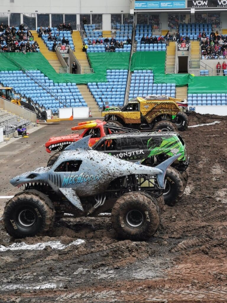 Three monster trucks, each with unique designs, are lined up on a muddy stadium track. The front truck resembles a shark and is labelled “Megalodon.” Behind it are two other trucks, one orange and one yellow, with a sparse crowd in blue stadium seats.
