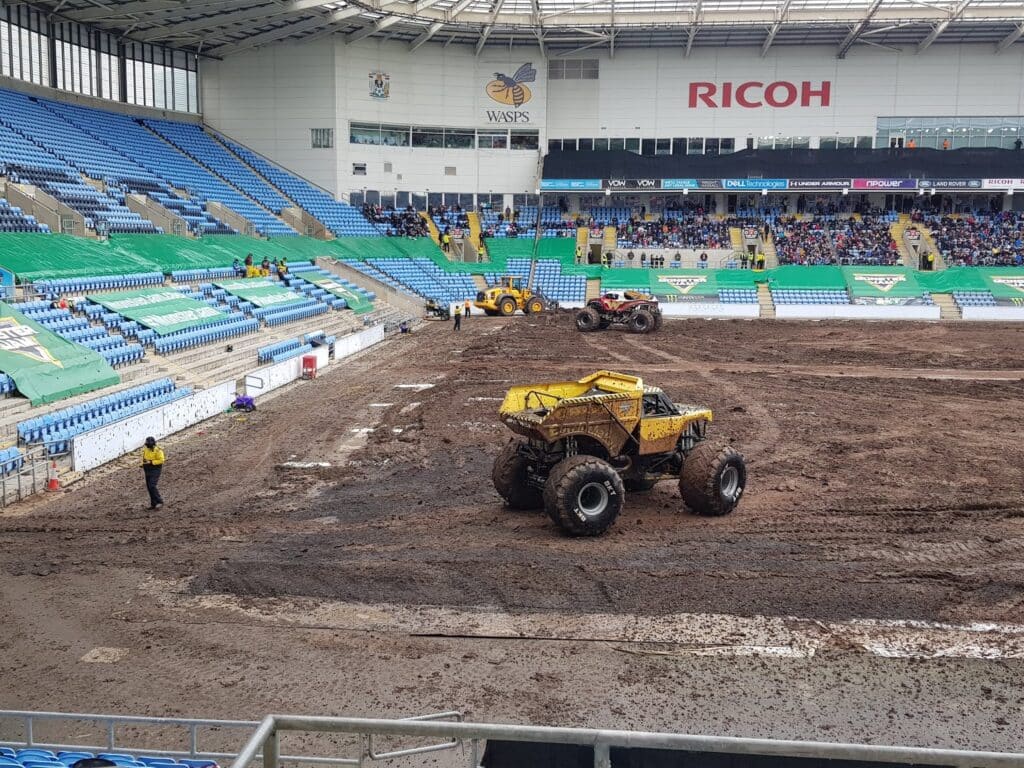 Large yellow monster lorries drive on a muddy arena inside a mostly empty stadium. Spectators sit in the background, some seats are vacant. A security guard stands near the track. Advertising banners and the Ricoh logo are visible on the stadium walls.