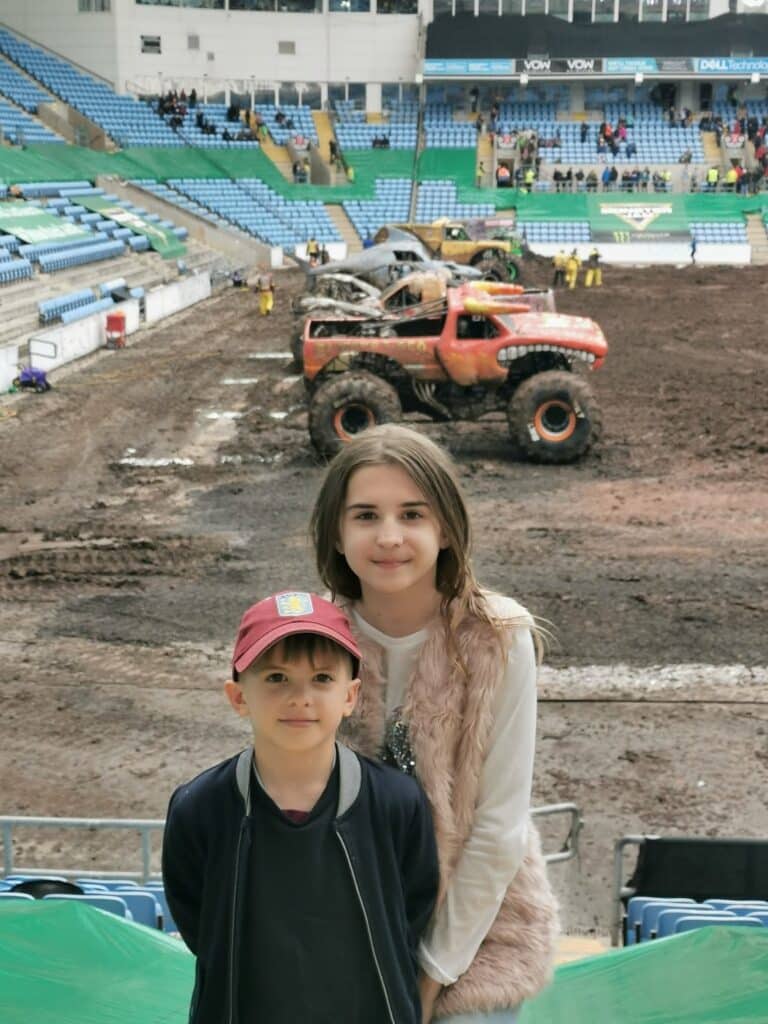 Two children, a boy in a red cap and a girl with long hair and a fur waistcoat, stand in a stadium with green and blue seats. Behind them, large muddy monster lorries are on a dirt arena, with spectators scattered in the background.