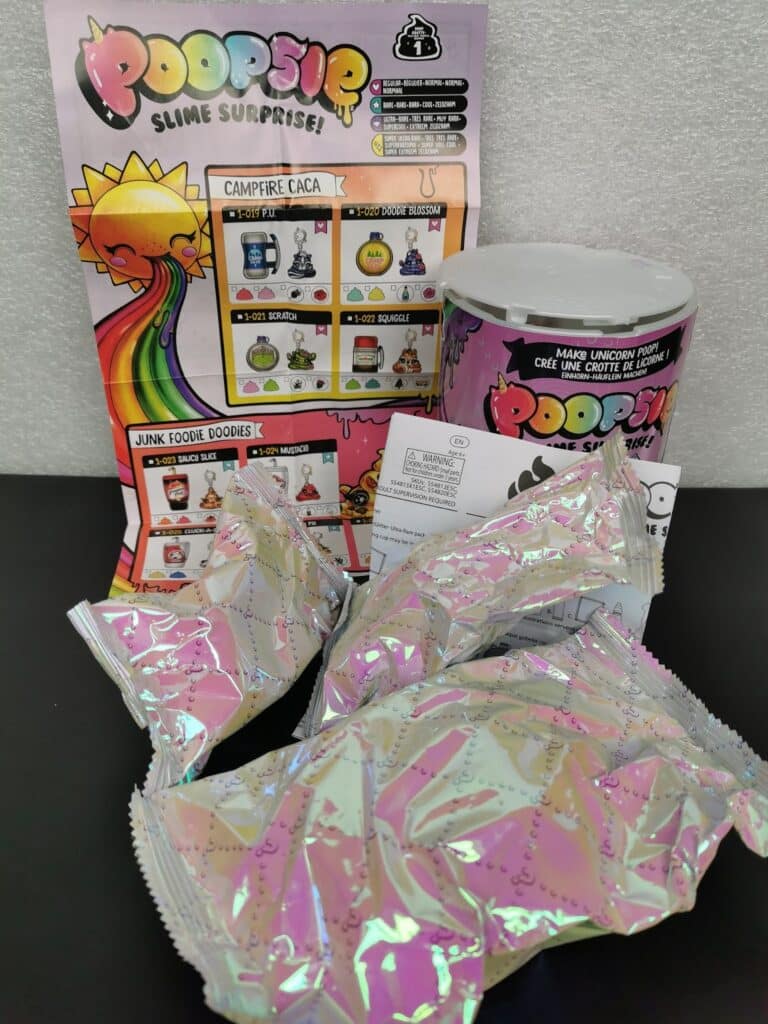 A Poopsie Slime Surprise toy set is displayed. Included are a colourful instruction sheet, a cylindrical container with playful graphics, several metallic, iridescent packets, and a small folded instruction paper, all on a black surface against a light background.