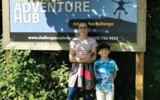Two children stand in front of a large "Baggeridge Adventure Hub" sign at Challenge Academy, surrounded by greenery. The older child wears glasses, a striped shirt, and black leggings, while the younger child wears a blue shirt with a nature pattern and white trousers.