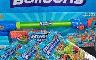 A Zuru Bunch O Balloons set includes a large blue and green water blaster and three packs of colourful self-sealing water balloons in blue, green, pink, and white. Packaging features playful graphics and images of children playing with water balloons.