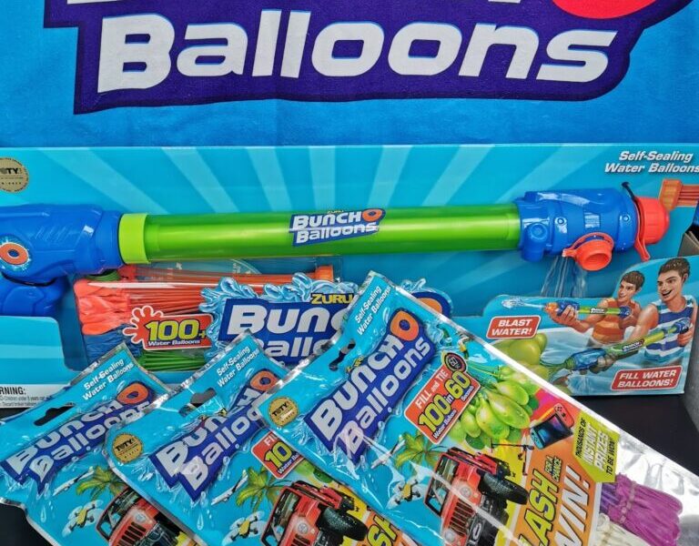 A Zuru Bunch O Balloons set includes a large blue and green water blaster and three packs of colourful self-sealing water balloons in blue, green, pink, and white. Packaging features playful graphics and images of children playing with water balloons.