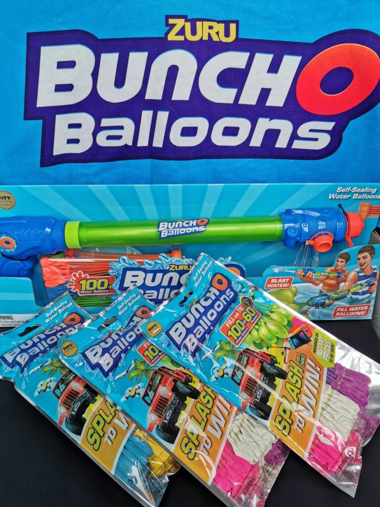 Zuru Bunch O Balloons packaging, featuring a blue box with a water blaster toy and bright text. In front, three packs of self-sealing water balloons in assorted colours (pink, white, green) are shown, labelled “Splash to Win!” with 100+ balloons per pack.