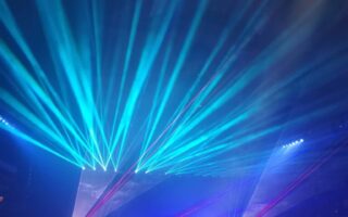 A concert stage with musicians is bathed in blue and purple lights, with bright laser beams shooting upward and outward, creating dynamic patterns above the audience. Silhouetted figures watch from the dark foreground, adding to the lively atmosphere.