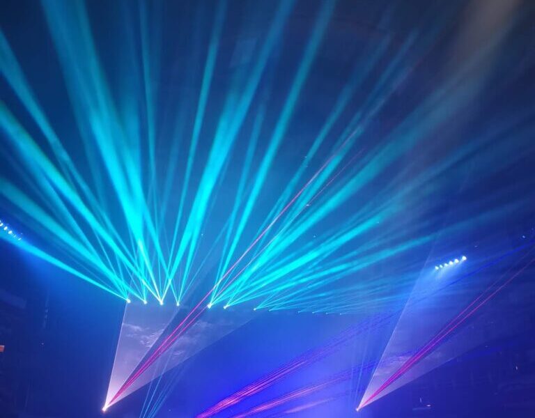 A concert stage with musicians is bathed in blue and purple lights, with bright laser beams shooting upward and outward, creating dynamic patterns above the audience. Silhouetted figures watch from the dark foreground, adding to the lively atmosphere.