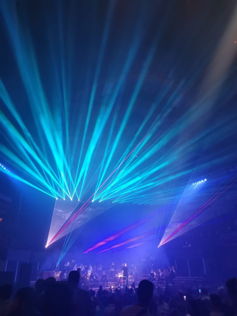 Blue and purple laser lights fan out above a concert stage, illuminating a hazy atmosphere. Musicians are visible on stage, and silhouettes of audience members can be seen in the foreground, watching the vibrant light show.